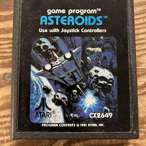 Puede incluir: Cartucho de videojuego Atari Asteroids negro. El cartucho presenta el título del juego en azul, un gráfico de nave espacial y el logotipo de Atari. El texto dice "Use with Joystick Controllers" y "Program Contents © 1981 Atari, Inc."