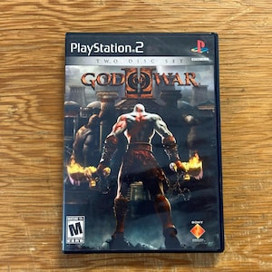 May include: A PlayStation 2 video game case for God of War II, a two-disc set. The cover art features a muscular, pale-skinned warrior standing with back to the viewer, holding two glowing blades, with the title God of War II and an M rating visible.