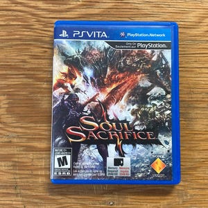 Peut inclure: Un boîtier de jeu bleu en plastique pour PlayStation Vita intitulé Soul Sacrifice. La jaquette présente une illustration fantastique sombre et détaillée avec un personnage central et un monstre. Le boîtier porte le logo PS Vita et l'étiquette M.