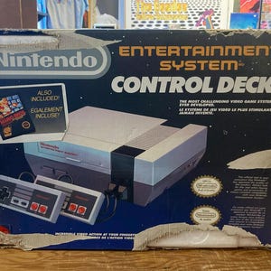 May include: A vintage Nintendo Entertainment System Control Deck box. The box is navy blue with white and orange text. The box includes the console, two controllers, and a game cartridge. The box is on a wooden surface.