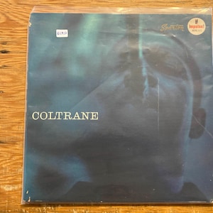 Może przedstawiać: Album winylowy zatytułowany "Coltrane" z abstrakcyjnym niebiesko-czarnym wzorem. Okładka albumu zawiera napis "Coltrane" białymi literami. Album jest w plastikowej folii i ma naklejkę w prawym górnym rogu.