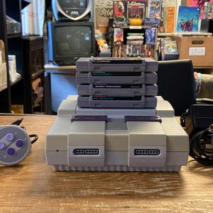 May include: A grey Super Nintendo game console with a matching controller, a stack of four grey game cartridges, and a black power adapter with a cord. The console features two controller ports on the front and a top-loading cartridge slot.