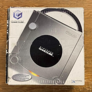 May include: A silver Nintendo GameCube console in its original box. The console has a circular disc tray and a black center with the GameCube logo. The box is white with the GameCube logo and text. "Limited Edition Platinum" is printed on the box.