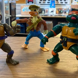 Op de afbeelding: Drie actiefiguren op een houten oppervlak. De figuren omvatten twee Teenage Mutant Ninja Turtles in groen en oranje, en een eendachtig personage in een bruine jas en een blauwe broek. De figuren staan in actieposities.