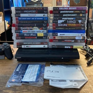 May include: A black Sony PlayStation 3 console with a wireless controller, a stack of twenty-two video game cases, power and AV cables, and two instruction manuals. The game titles include Uncharted 2, Assassin's Creed, Payday 2, and Mafia II.