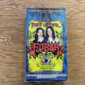 Puede incluir: Una cinta VHS de la película "Fubar" con un diseño de chaqueta vaquera. La cinta presenta a dos hombres con pelo largo y el texto "JUST GIVE'R" y "FUBAR". El texto adicional incluye "Le meilleur film de rock'n'roll depuis des années!"