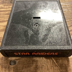 Peut inclure: Une cartouche de jeu vidéo rectangulaire noire avec les mots "STAR RAIDERS" en lettres orange. La cartouche a une petite fente et une vis sur le dessus. La surface a un aspect usé et texturé.