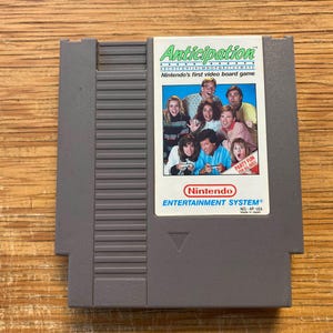 May include: A grey Nintendo Entertainment System game cartridge for the game "Anticipation." The label features a group of people and the text "Nintendo's first video board game." The cartridge is on a wooden surface.