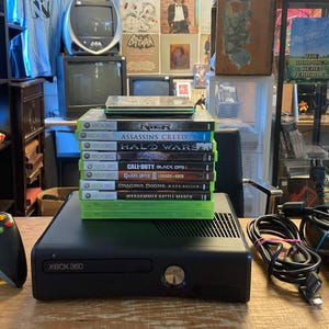 May include: A black Xbox 360 game console with a matching black wireless controller, a power brick with attached cables, and a vertical stack of eight video games including titles like Nier, Assassin's Creed, Halo Wars, and Call of Duty Black Ops II.
