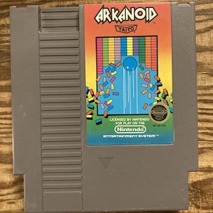May include: A grey Nintendo Entertainment System game cartridge for Arkanoid. The label features the game's title in orange and a colourful graphic of a waterfall and geometric shapes. The Nintendo logo is visible.