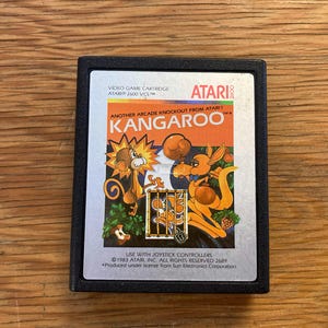 Peut inclure: Cartouche de jeu vidéo Atari 2600 pour le jeu "Kangaroo". L'étiquette présente un kangourou et un singe de dessin animé dans un design coloré. La cartouche est dans un boîtier en plastique noir.