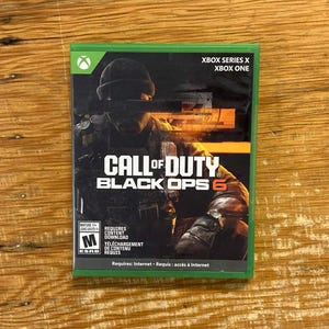Puede incluir: Una caja de videojuego Call of Duty Black Ops 6 para Xbox Series X y Xbox One. La portada muestra a un soldado con gorro y una barra negra sobre los ojos, el título en blanco y naranja, y una clasificación ESRB M para público adulto.