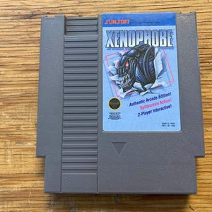 May include: A grey Xenophobe video game cartridge for the Nintendo Entertainment System. The label shows a menacing alien bursting through a wall, with text that reads "Authentic Arcade Edition!" and "2-Player Interactive!"