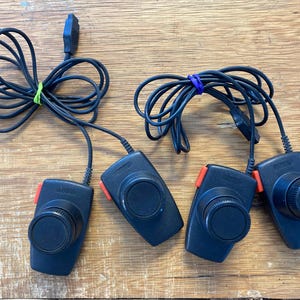 Puede incluir: Cuatro mandos de videojuegos negros con detalles rojos y un dial grande. Los mandos están conectados a cables negros, agrupados con una goma elástica verde y morada. Los mandos están sobre una superficie de madera.