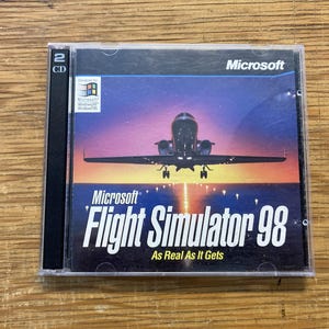 May include: Microsoft Flight Simulator 98 CD-ROM case. The cover features a jet plane taking off against a sunset backdrop. The game title and "As Real As It Gets" are prominently displayed.