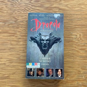 Puede incluir: Una cinta VHS de la película "Drácula" de Francis Ford Coppola. La portada presenta una ilustración oscura de una cara demoníaca y el título en escritura roja. También se ve el texto "L'AMOUR NE MEURT JAMAIS".