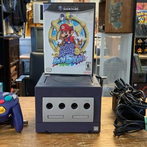 Peut inclure: Une console Nintendo GameCube violette avec une manette violette assortie, un câble d'alimentation noir et un boîtier de jeu Super Mario Sunshine. La console présente un panneau avant argenté avec quatre ports manette et deux emplacements carte mémoire.