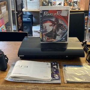 May include: A black Sony PlayStation 3 console with a game case for "Prince of Persia" on top. A black controller, cables, and manuals are also on the wooden surface. The game console is a classic gaming item.
