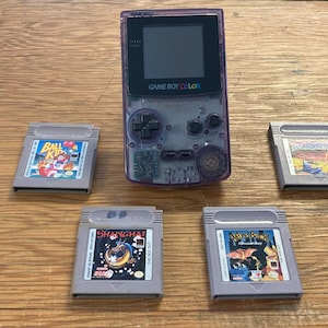 Puede incluir: Una consola de juegos portátil Game Boy Color de color morado translúcido con un marco de pantalla negro y cuatro cartuchos de juego grises colocados alrededor. Los cartuchos tienen etiquetas para los juegos Ball Kid, Shanghai y otros dos títulos.