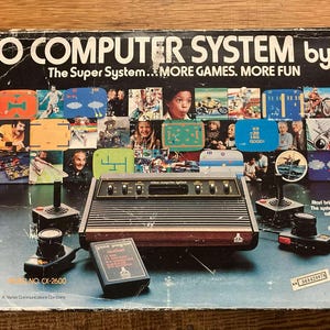 Può includere: Scatola del sistema video Atari vintage con la scritta "VIDEO COMPUTER SYSTEM by ATARI". La scatola presenta immagini di giochi e persone. Include una console, joystick e una cartuccia di gioco.
