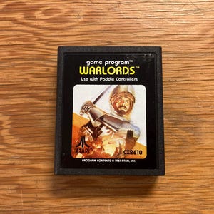 Peut inclure: Cartouche de jeu Atari Warlords noire. L'étiquette présente une illustration de chevalier avec le texte "WARLORDS" et "Use with Paddle Controllers." La cartouche est étiquetée "CX2610" et comprend le logo Atari.