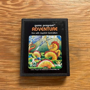 Puede incluir: Cartucho de videojuego Atari Adventure negro. La etiqueta presenta una colorida ilustración de un dragón y un castillo. El texto en la etiqueta dice "ADVENTURE" y "Use with Joystick Controllers".