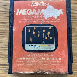 Puede incluir: Un cartucho de videojuego Atari vintage para el juego "Megamania". El cartucho tiene una carcasa de plástico negro con una etiqueta naranja. La etiqueta presenta el título del juego, un gráfico de pantalla de juego retro y el logotipo de Activision.