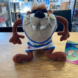 May include: A plush Taz the Tasmanian Devil toy wearing a white and blue basketball uniform. The toy has brown fur, large white teeth, and a black nose. A tag with a cartoon image of Taz and a basketball is next to the toy.