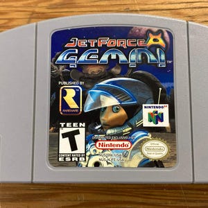 Peut inclure: Une cartouche de jeu vidéo grise pour Nintendo 64 intitulée Jet Force Gemini. L'étiquette présente un personnage en combinaison spatiale bleue et blanche, le logo Rareware, le logo Nintendo 64 et le symbole de classification ESRB Teen.