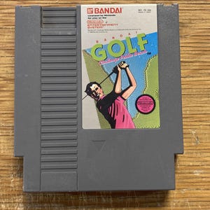 May include: A grey Nintendo Entertainment System (NES) cartridge for the game "Bandai Golf: Challenge Pebble Beach." The label shows a golfer in a pink shirt, with the game's title in green and blue.