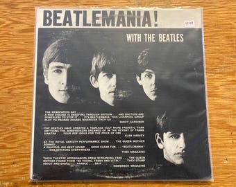 May include: Vintage vinyl record album cover for "Beatlemania! With The Beatles." The black and white cover features four band member portraits and text. The album is in a plastic sleeve and is on a wooden surface.