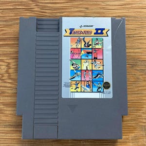 Könnte beinhalten: Eine graue, rechteckige NES-Videospielkassette für Track & Field II von Konami. Das Etikett zeigt ein Raster aus farbenfrohen Illustrationen verschiedener olympischer Sportarten, darunter Bogenschießen, Fechten und Gymnastik, mit dem Spieletitel.