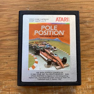 Pode incluir: Cartucho de videogame Atari 2600 para Pole Position. O rótulo apresenta um carro de corrida em uma pista com o título do jogo em letras grandes laranja. O cartucho é preto.