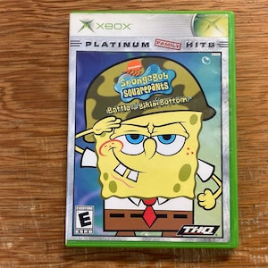 Può includere: Una custodia per videogioco per la console Xbox con il titolo SpongeBob SquarePants: Battle for Bikini Bottom. La copertina mostra un personaggio giallo che indossa un elmetto militare mimetico e fa il saluto, con l'etichetta Platinum Hits.