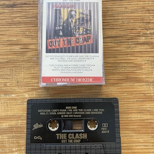以下が含まれることがあります： 「The Clash - Cut the Crap」アルバムのカセットテープ2本。上のカセットはアルバムアート付きのクリアケースに入っています。下のカセットは黒色で、アルバムタイトルと曲リストを含む白い文字が書かれています。