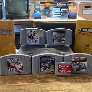 May include: A grey Nintendo 64 game console bundle including a grey controller, five game cartridges, a red Memory Card Plus, two yellow instruction manuals for Star Wars Episode I Racer and F-1 World Grand Prix, and black power and AV cables.