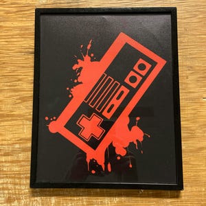 May include: Framed art print featuring a red graphic of a classic video game controller on a black background. The controller design is slightly angled with red paint splatter accents. The frame is black.