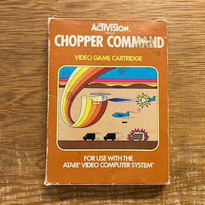 Op de afbeelding: Een vintage Atari Chopper Command videogamecartridge. De oranje doos heeft een kleurrijke illustratie van een helikopter, vliegtuigen en explosies. De tekst op de doos luidt "ACTIVISION" en "CHOPPER COMMAND".