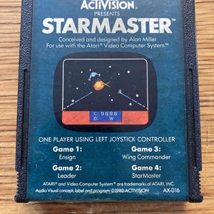 May include: A vintage Atari video game cartridge for Starmaster. The cartridge is dark blue with white text, including the game title and "Activision Presents." The game screen shows a spaceship and other graphics. The cartridge also lists the game's four modes.