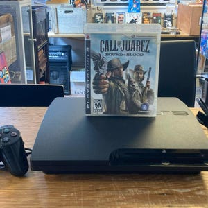 May include: A black PlayStation 3 console with a game case for "Call of Juarez: Bound in Blood" and a black controller. A black power cable is also present. The items are on a wooden table.