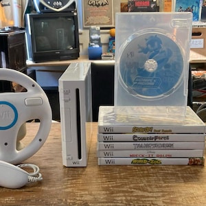 Könnte beinhalten: Ein weißes Nintendo Wii-Spielkonsolen-Set bestehend aus einem Lenkrad-Controller, einer Fernbedienung, einem Nunchuk, einem Netzteil und einem Stapel von fünf Videospielen mit einer sichtbaren Disc in einer transparenten Hülle auf einer Holzfläche.
