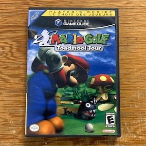 Może przedstawiać: Etui na grę wideo Nintendo GameCube Mario Golf Toadstool Tour. Okładka przedstawia Mario zamachującego się kijem golfowym na zielonym polu, obok postaci Chain Chomp i dużego grzyba. Na górze znajduje się etykieta Player's Choice.