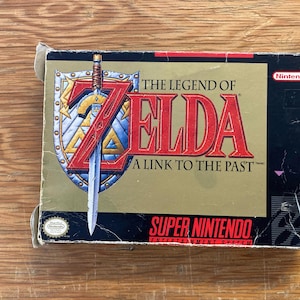 May include: The Legend of Zelda: A Link to the Past Super Nintendo game box. The box features a gold background with the game title in red and a sword. The Nintendo logo is in the upper right corner.