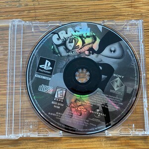 May include: A PlayStation game disc for Crash Bandicoot 2: Cortex Strikes Back, in a clear plastic case. The disc features the game's title and character artwork. The case is open, revealing the disc on a wooden surface.