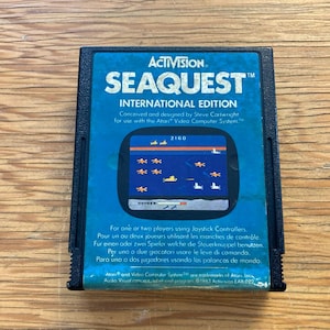 Può includere: Una cartuccia per videogiochi nera Activision Seaquest International Edition. L'etichetta blu presenta il titolo del gioco, una grafica dello schermo di gioco e testo in più lingue. La cartuccia è per l'Atari Video Computer System.
