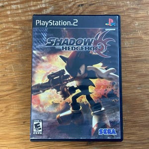 May include: A PlayStation 2 video game case for Shadow the Hedgehog. The cover art features the black character Shadow holding a firearm against an explosive background. The title Shadow the Hedgehog is printed in white and red, with the Sega logo below.