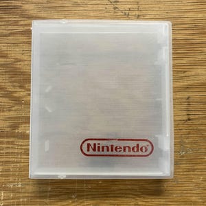 以下が含まれることがあります： 透明な長方形の任天堂ゲームカートリッジケース。ケースの右下隅には赤い任天堂のロゴがあります。ケースは半透明のプラスチック素材で作られています。
