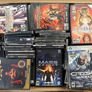 May include: A collection of various PC video game cases packed inside a cardboard box. Visible titles include Phantasy Star Universe, Dungeon Siege II, Fable The Lost Chapters, Diablo, Mass Effect, and Crysis Warhead, alongside several other game discs.