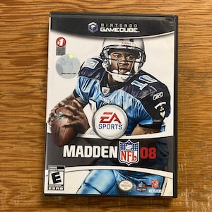 May include: A Nintendo GameCube video game case for Madden NFL 08. The cover features a football player in a blue and black uniform holding a football. The EA Sports logo, the NFL logo, and an ESRB E rating for Everyone are clearly visible on the front.