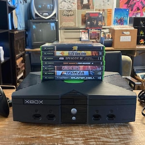 Puede incluir: Una consola de videojuegos Xbox original de color negro con un mando negro a juego, un conjunto de cables AV negros y una pila de seis cajas de videojuegos que incluyen títulos como Black Hawk Down, Episode III, Pro Skater 4 y Splinter Cell.